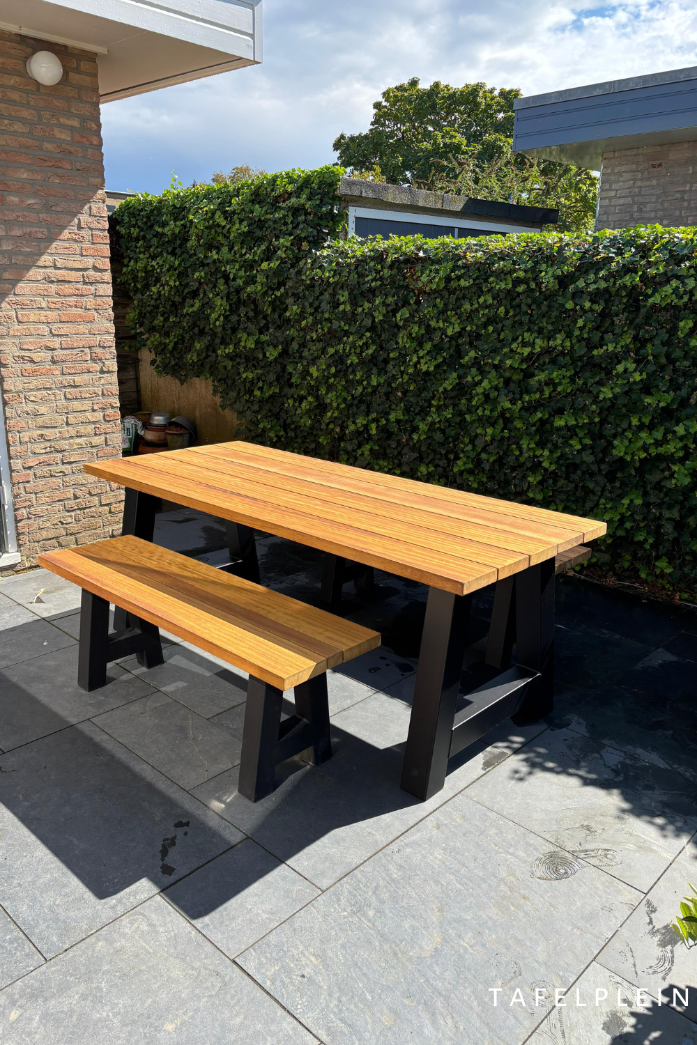 Tuintafel van iroko hout en stalen zwart gepoedercoate A poten in tuin op grijze tegels met bankje van iroko hout en zwart gepoedercoate A poten