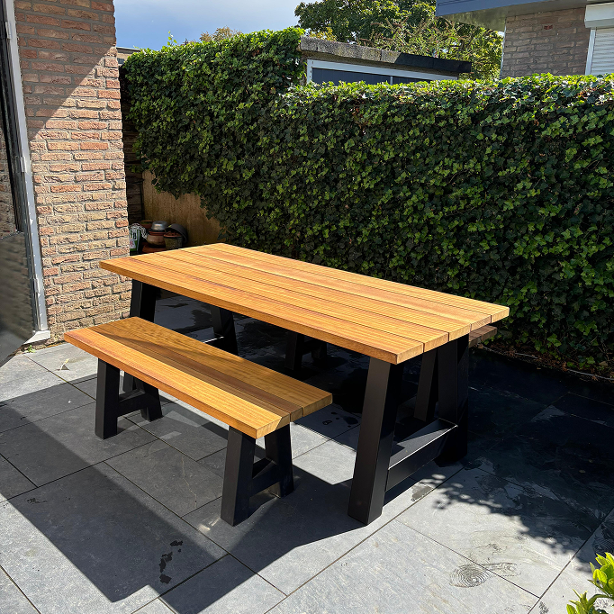Tuintafel van iroko hout en stalen zwart gepoedercoate A poten in tuin op grijze tegels met bankje van iroko hout en zwart gepoedercoate A poten